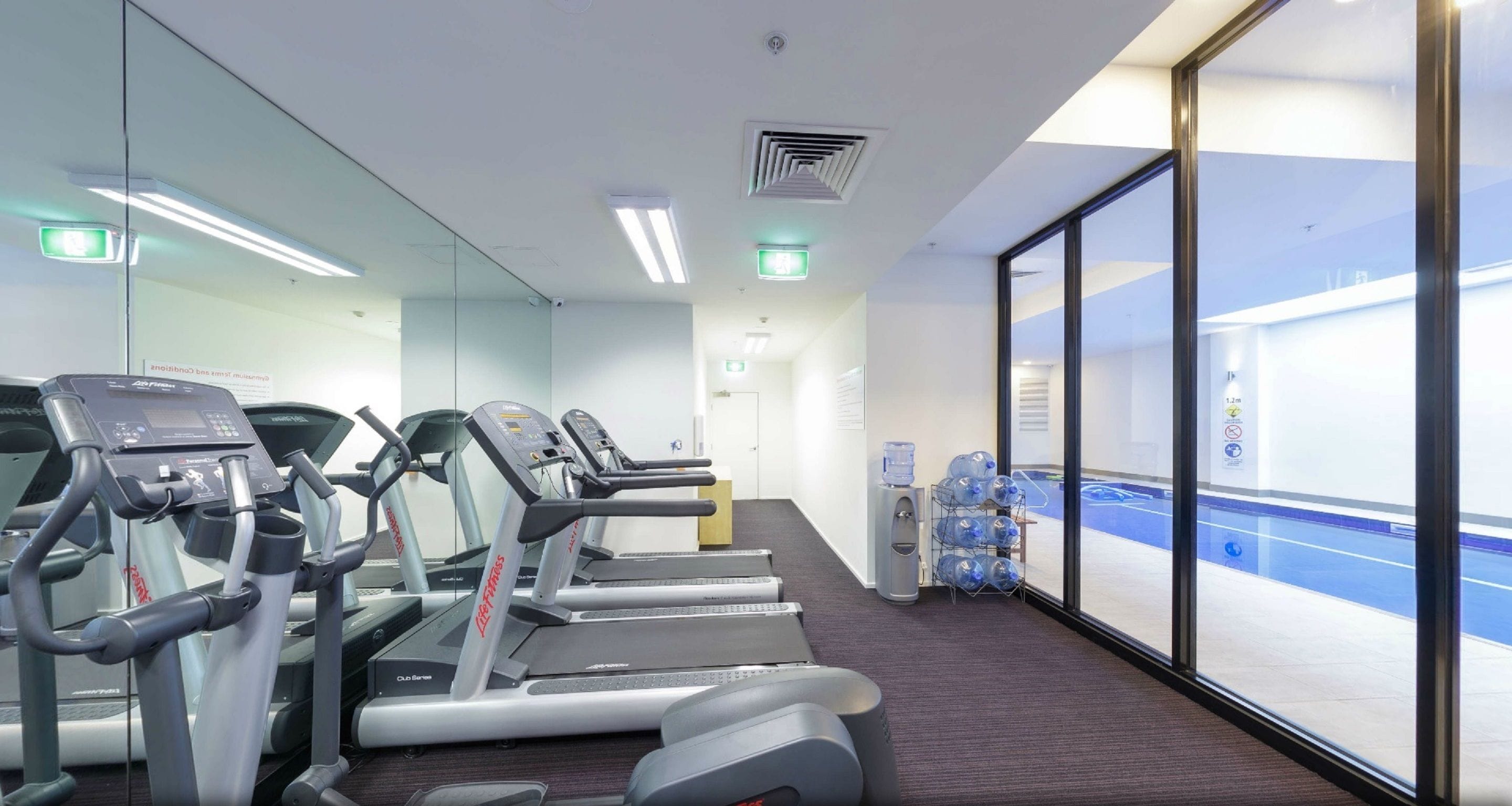 A gym with treadmills, a water dispenser, and a view of a pool through large windows.