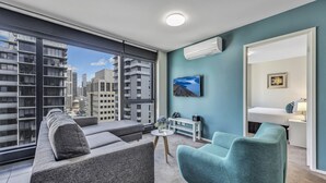 A modern living room with a sofa, armchairs, and a small table with a vase of flowers. A painting is on the wall, and there is a window with a view of tall buildings. - Pegasus Apart'Hotel (Melbourne)