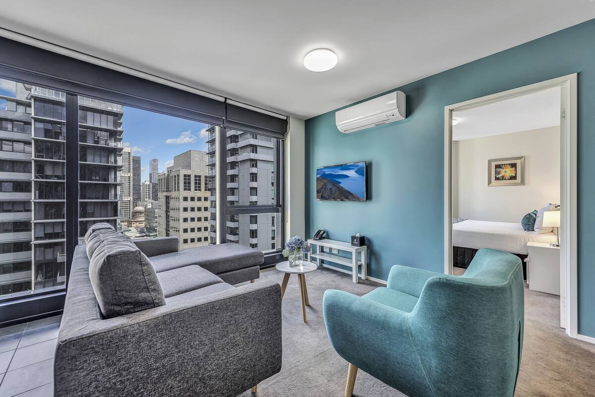 A modern living room with a sofa, armchairs, and a small table with a vase of flowers. A painting is on the wall, and there is a window with a view of tall buildings.