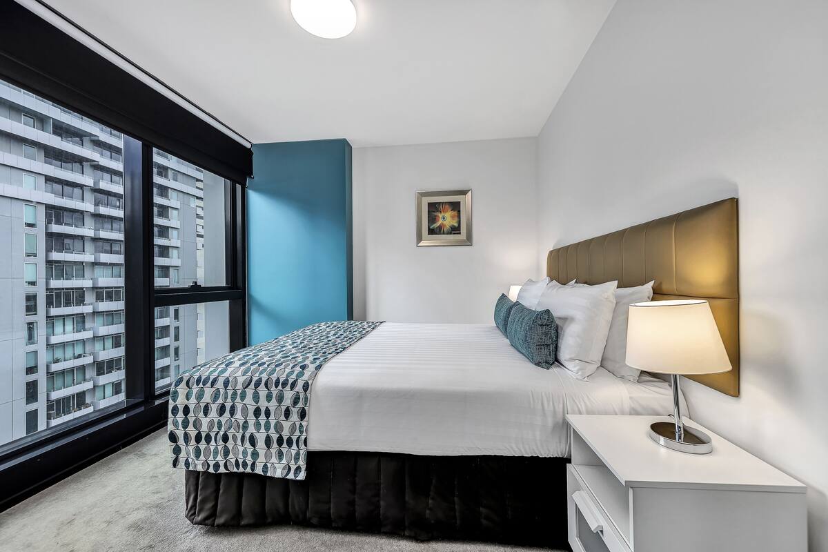 A modern hotel room with a large window, a bed with a patterned bedspread, a bedside lamp, and a framed picture on the wall.