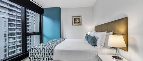 Una habitación de hotel moderna con un ventanal grande, una cama con colcha estampada, una lámpara de noche y un cuadro enmarcado en la pared.