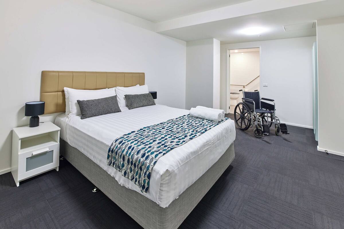 A hotel room with a large bed, a bedside table with a lamp, a wheelchair, and a door leading to a staircase.