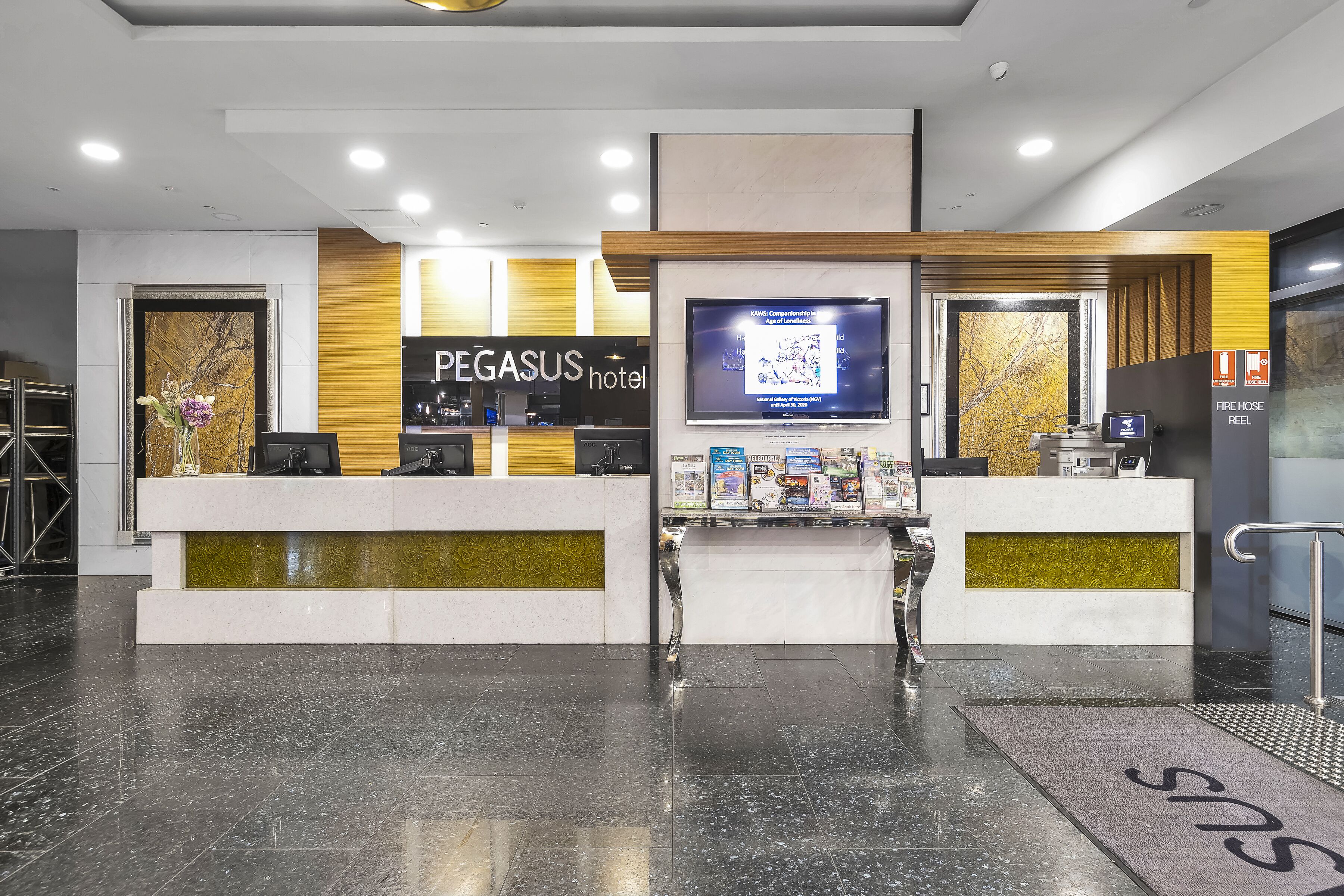 Uma área de recepção com piso de mármore, balcão de atendimento, tela de televisão e placa do Hotel Pegasus.