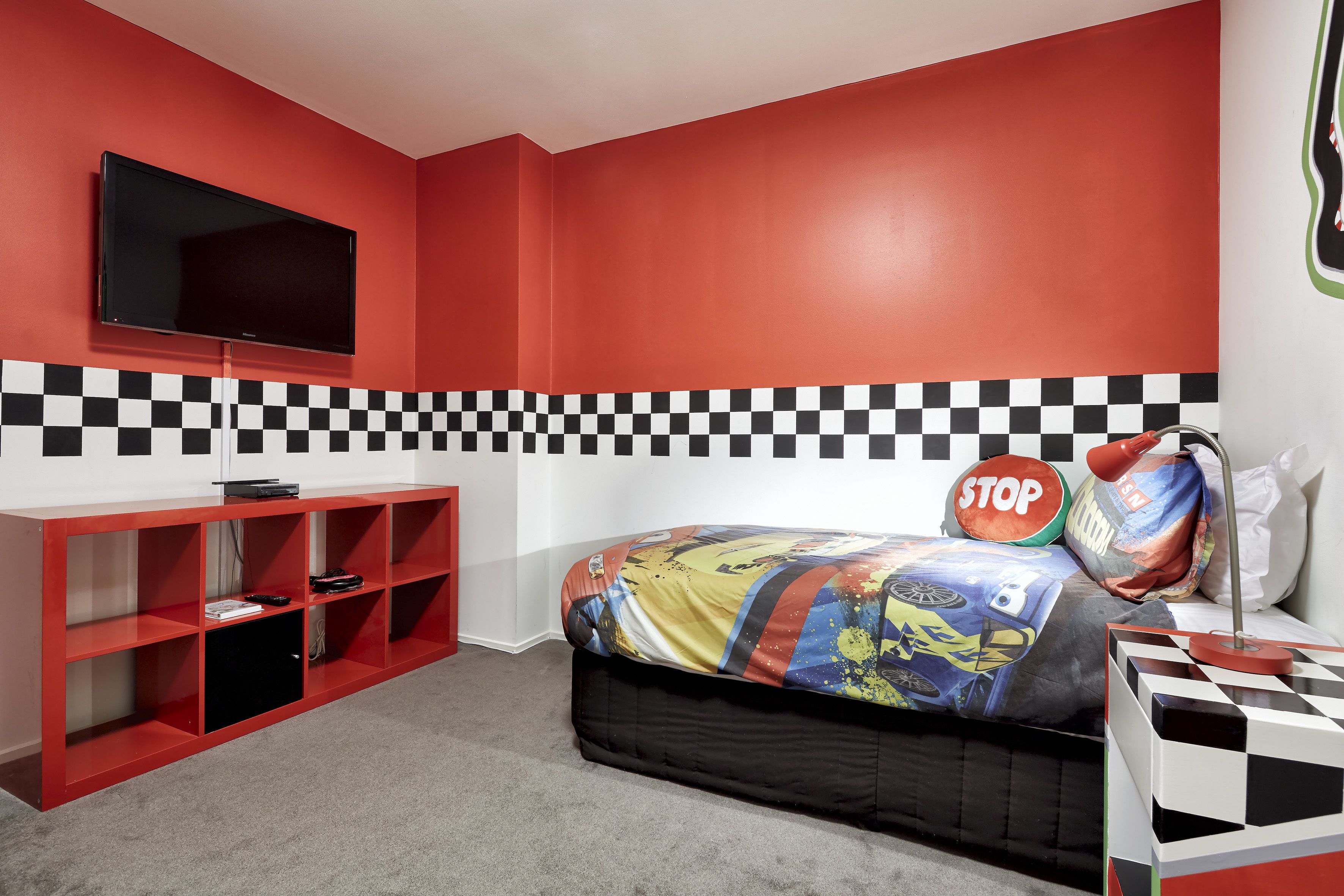 Une chambre avec un mur à carreaux, un lit recouvert d’une couverture colorée, un meuble TV rouge et un coussin orné du mot « STOP ».