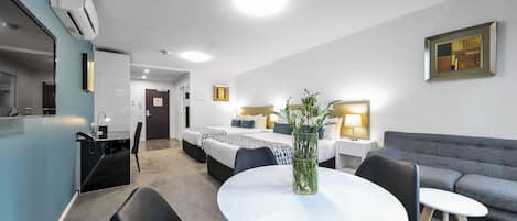 A modern hotel room with a kitchenette, a dining area with a round table, a sofa, and a bed with a headboard.