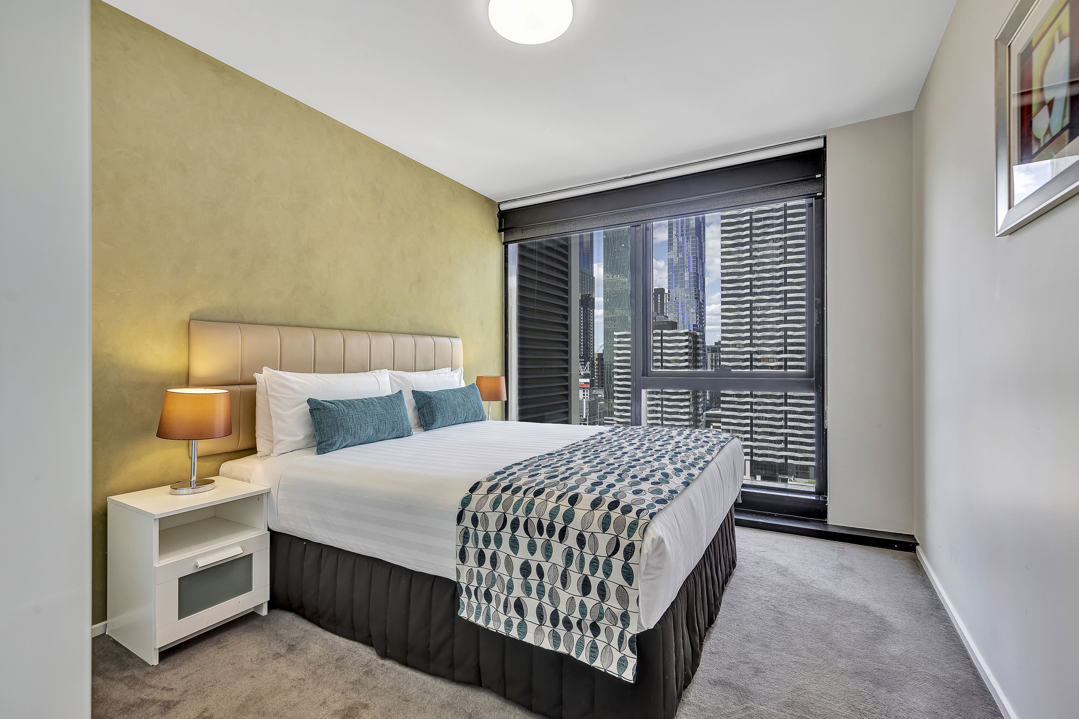Ein modernes Hotelzimmer mit einem großen Bett, einem Nachttisch, einer Lampe und Blick auf die Stadt durch ein großes Fenster.