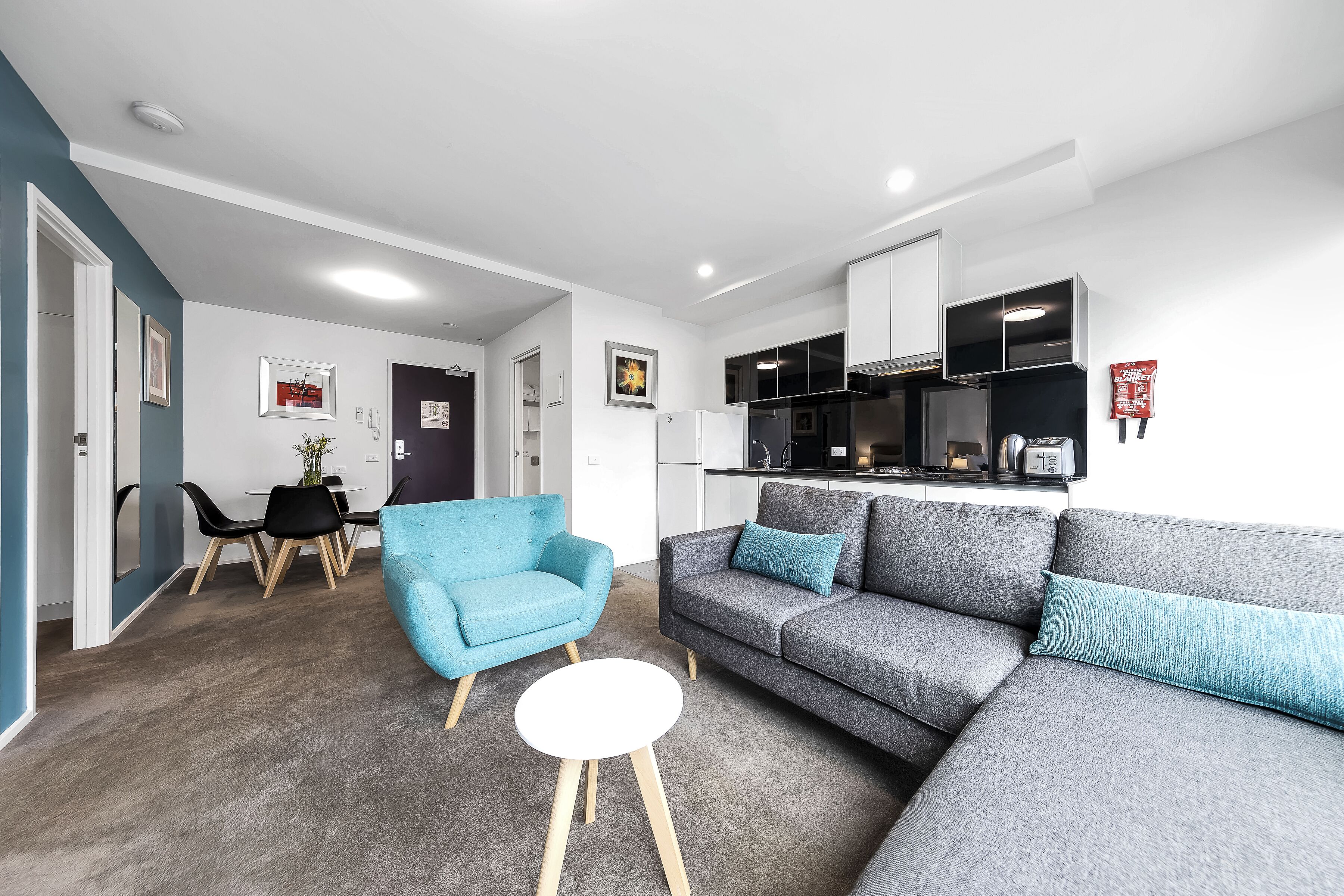 Una sala moderna con un sofá gris, un sillón azul y una pequeña mesa blanca. Al fondo, un área de comedor con una mesa y sillas.