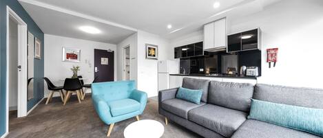 Una sala moderna con un sofá gris, un sillón azul y una pequeña mesa blanca. Al fondo, un área de comedor con una mesa y sillas.