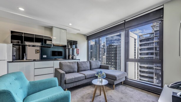 A modern apartment with a grey sofa, teal armchair, and a small round table with a vase of flowers. A kitchen with white cabinets and a large window offering a city view is visible. - Pegasus Apart'Hotel (Melbourne)