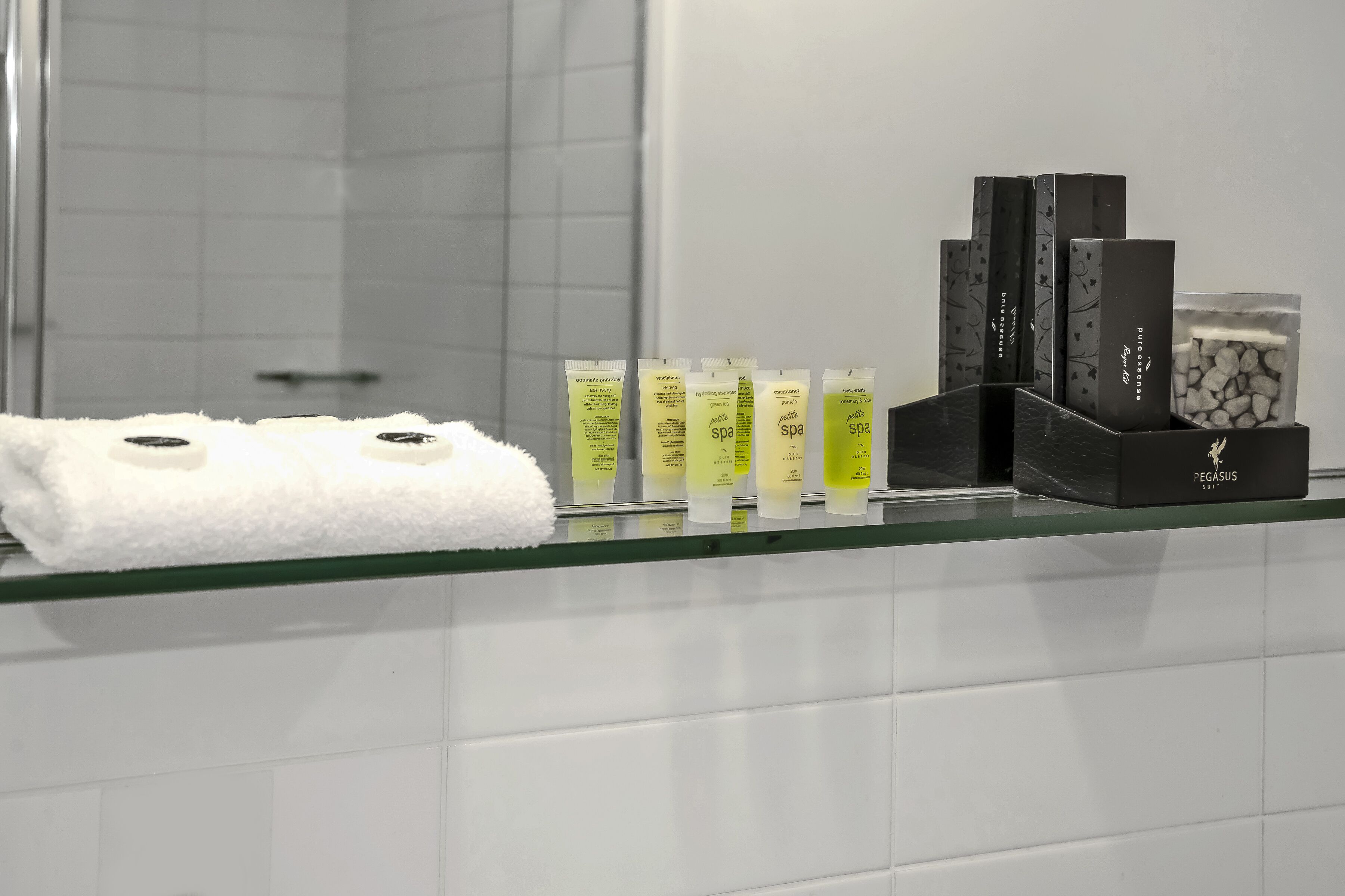 A bathroom with a glass shower enclosure, white tiles, and a glass shelf holding toiletries and a white towel.