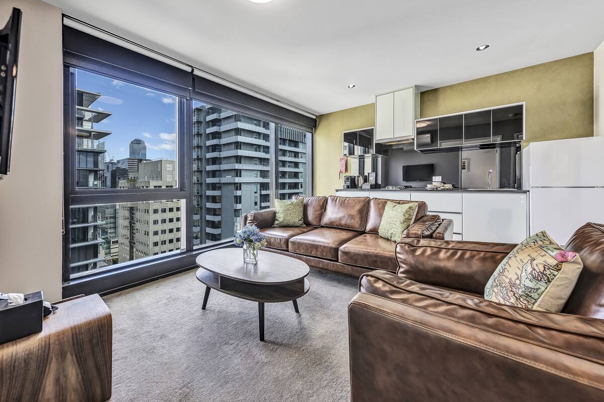 A modern apartment with a large window, a brown leather sofa, a small coffee table, and a kitchen area.