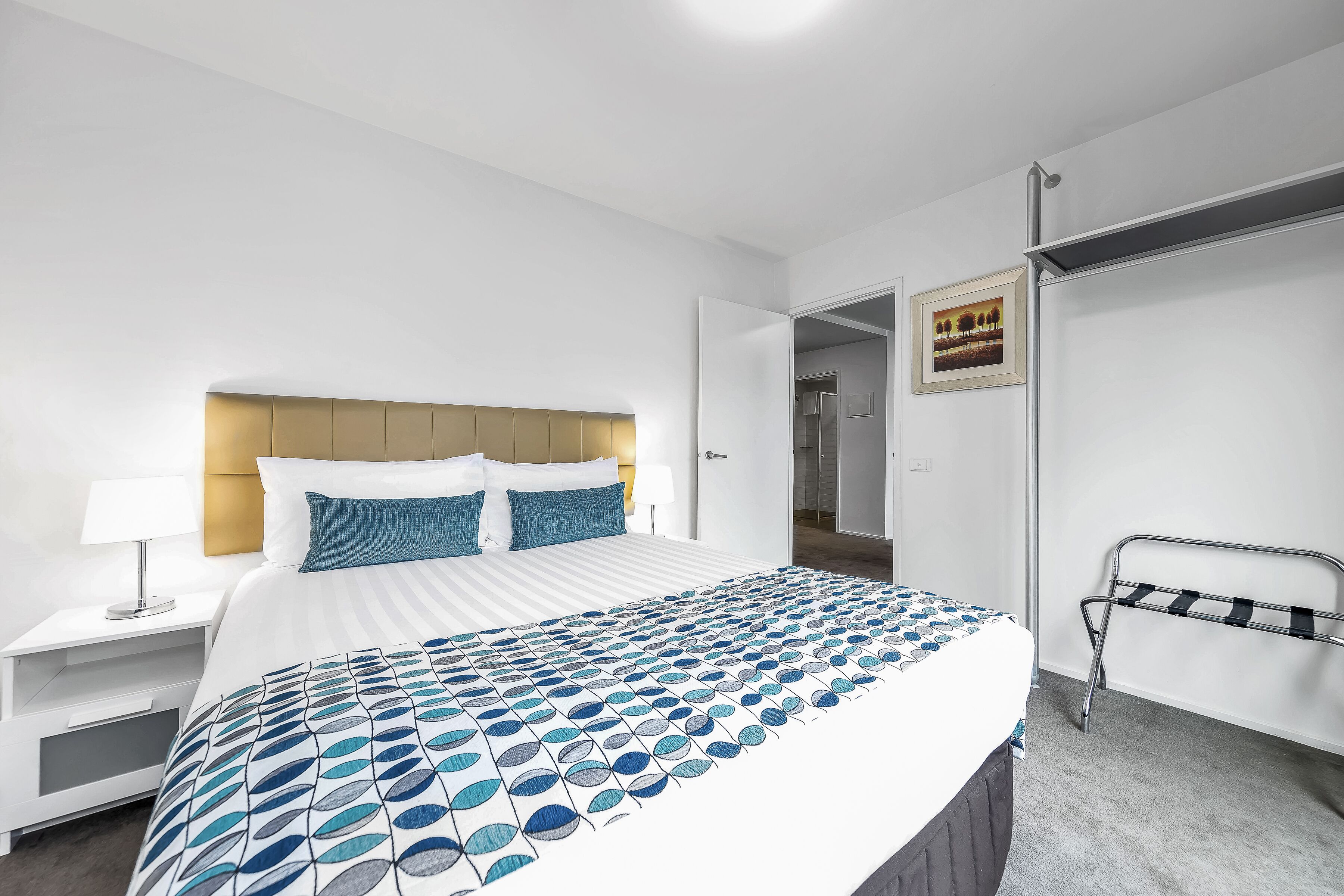 A hotel room with a neatly made bed, a bedside table with a lamp, and a metal rack against the wall.