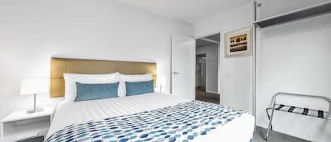 A hotel room with a neatly made bed, a bedside table with a lamp, and a metal rack against the wall.