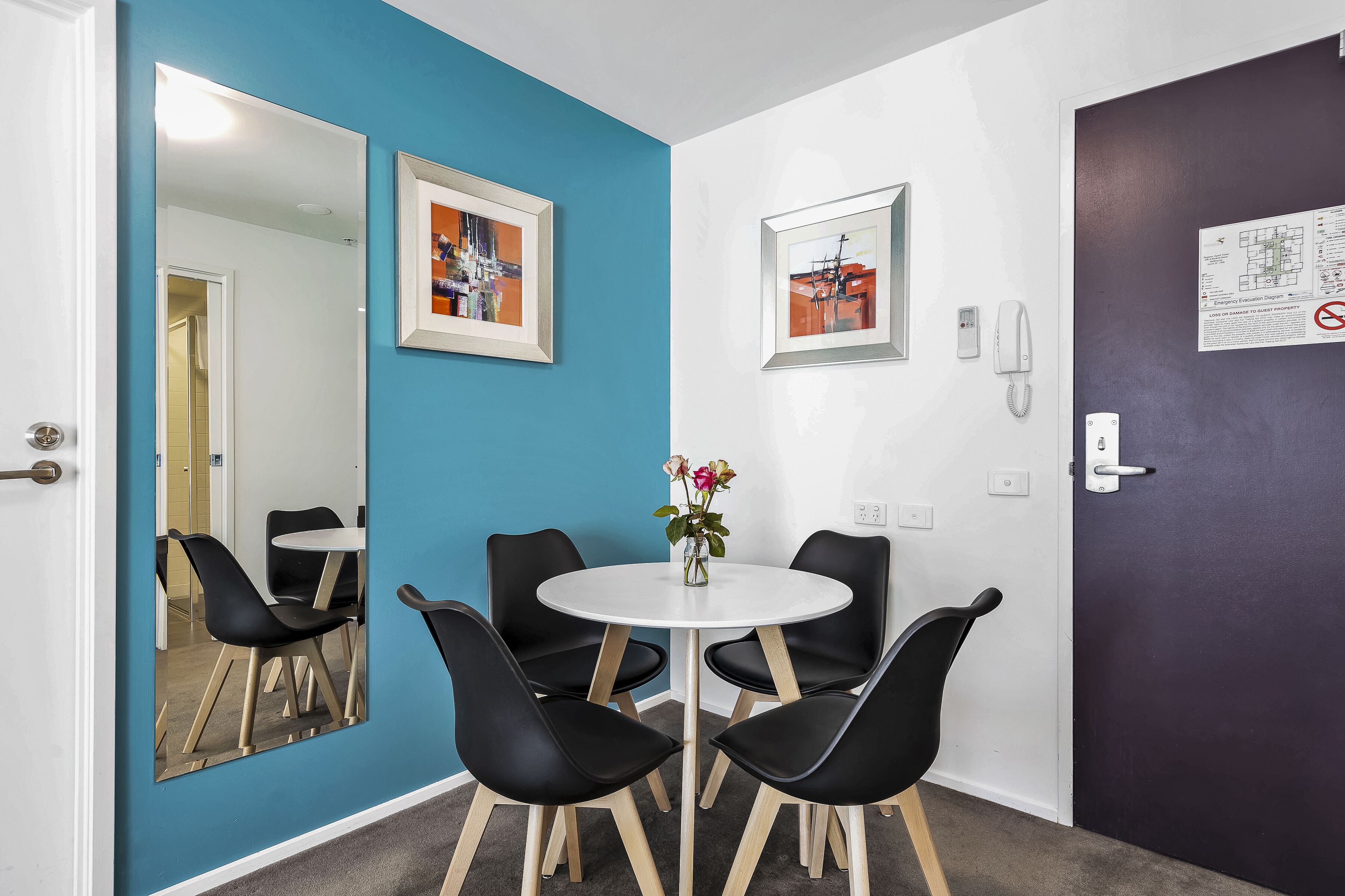 A room with a blue wall, a white door, a round table with four black chairs, and a mirror reflecting the room.