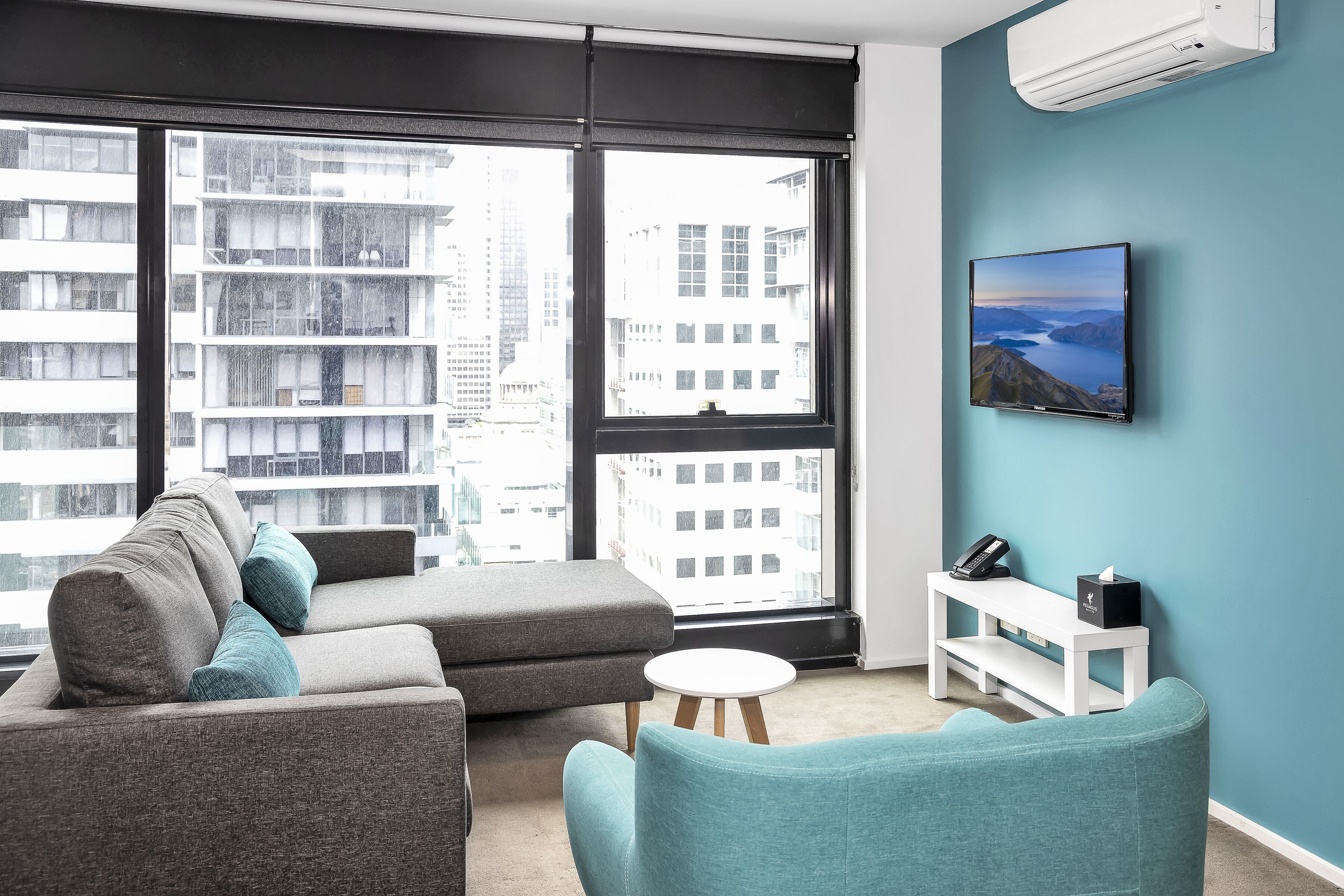 Una sala de estar moderna con un sofá seccional gris, un sillón color turquesa y una mesita blanca. Un televisor de pantalla plana está montado en la pared, y un banco blanco con un teléfono y pañuelos está apoyado contra la pared.