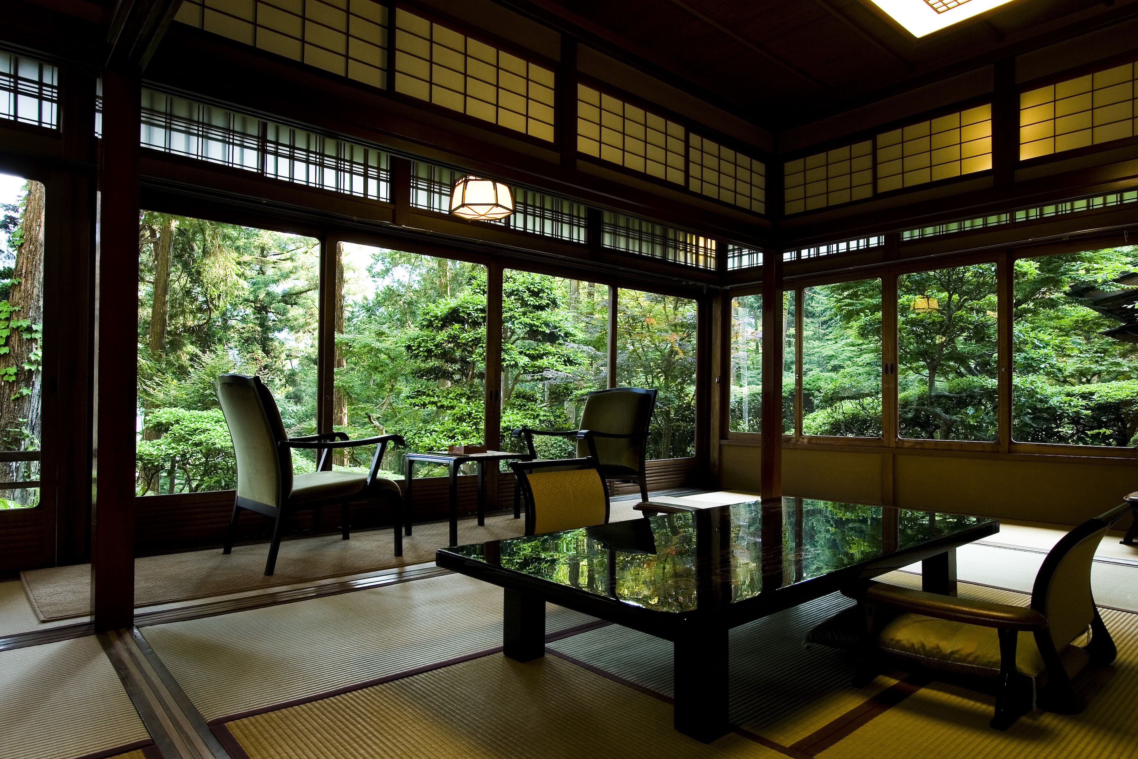 Um quarto japonês tradicional com tatames, uma mesa baixa e cadeiras. Grandes janelas revelam uma vista de vegetação exuberante lá fora.