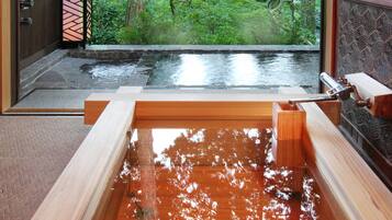 A wooden hot tub with a stone surround, a wooden deck, and a view of a garden with a water feature.