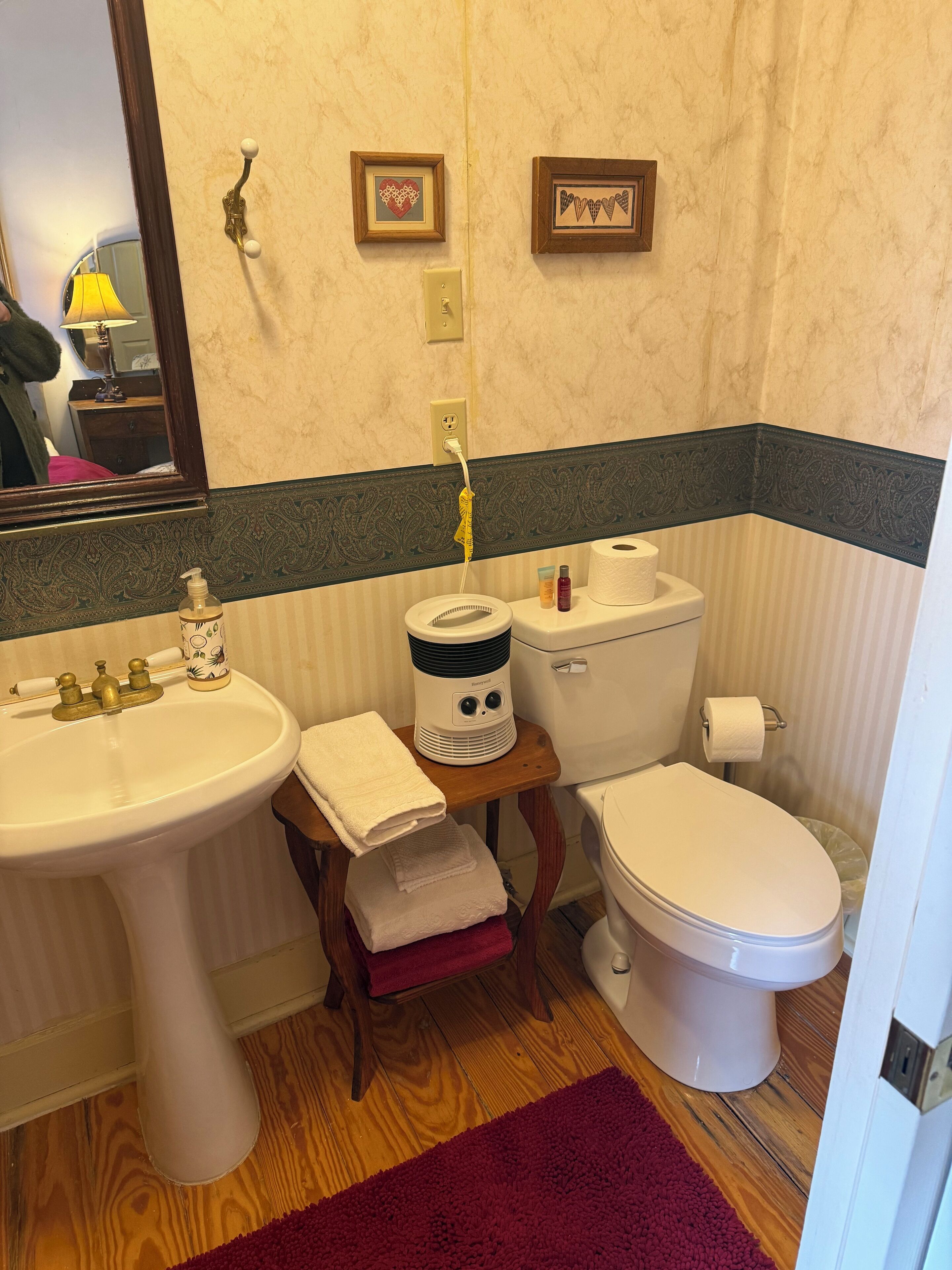 A bathroom with a sink, toilet, and a small table with a heater. There are towels on the floor and a mirror above the sink.