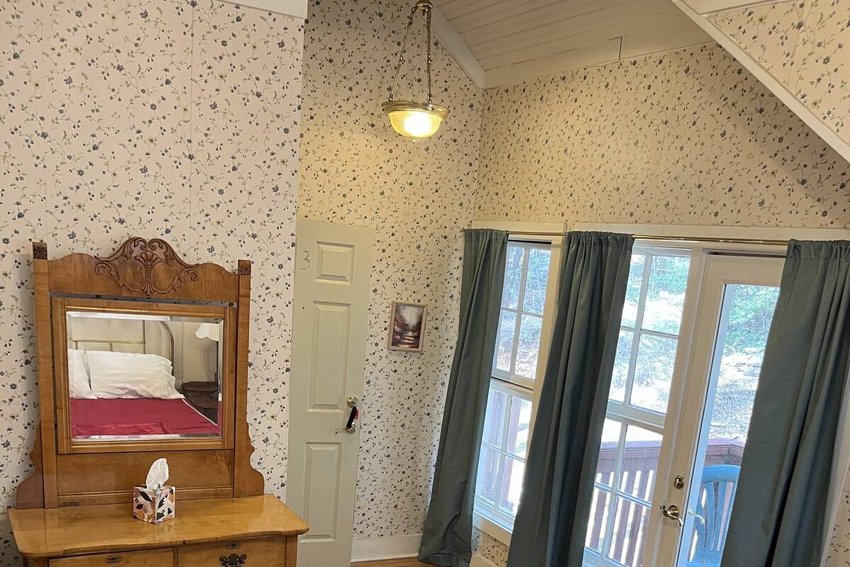 A bedroom with a wooden dresser, a bed with a red bedspread, a mirror, a door with a picture, and a view through a window with curtains.