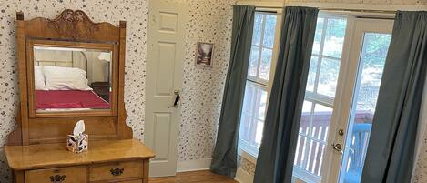 Een slaapkamer met een houten dressoir, een bed met een rode sprei, een spiegel, een deur met een afbeelding en uitzicht door een raam met gordijnen.