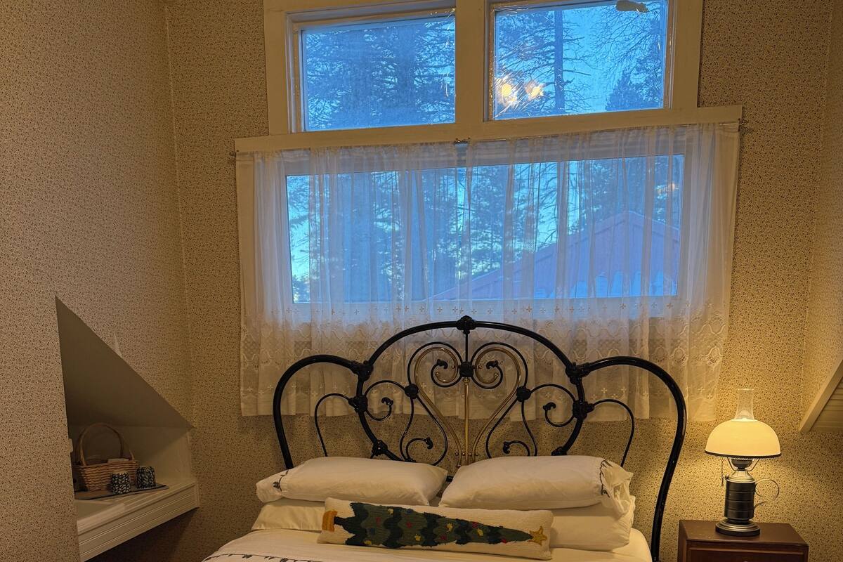 A bedroom with a bed, a nightstand with a lamp, a window with curtains, and a shelf with a basket.