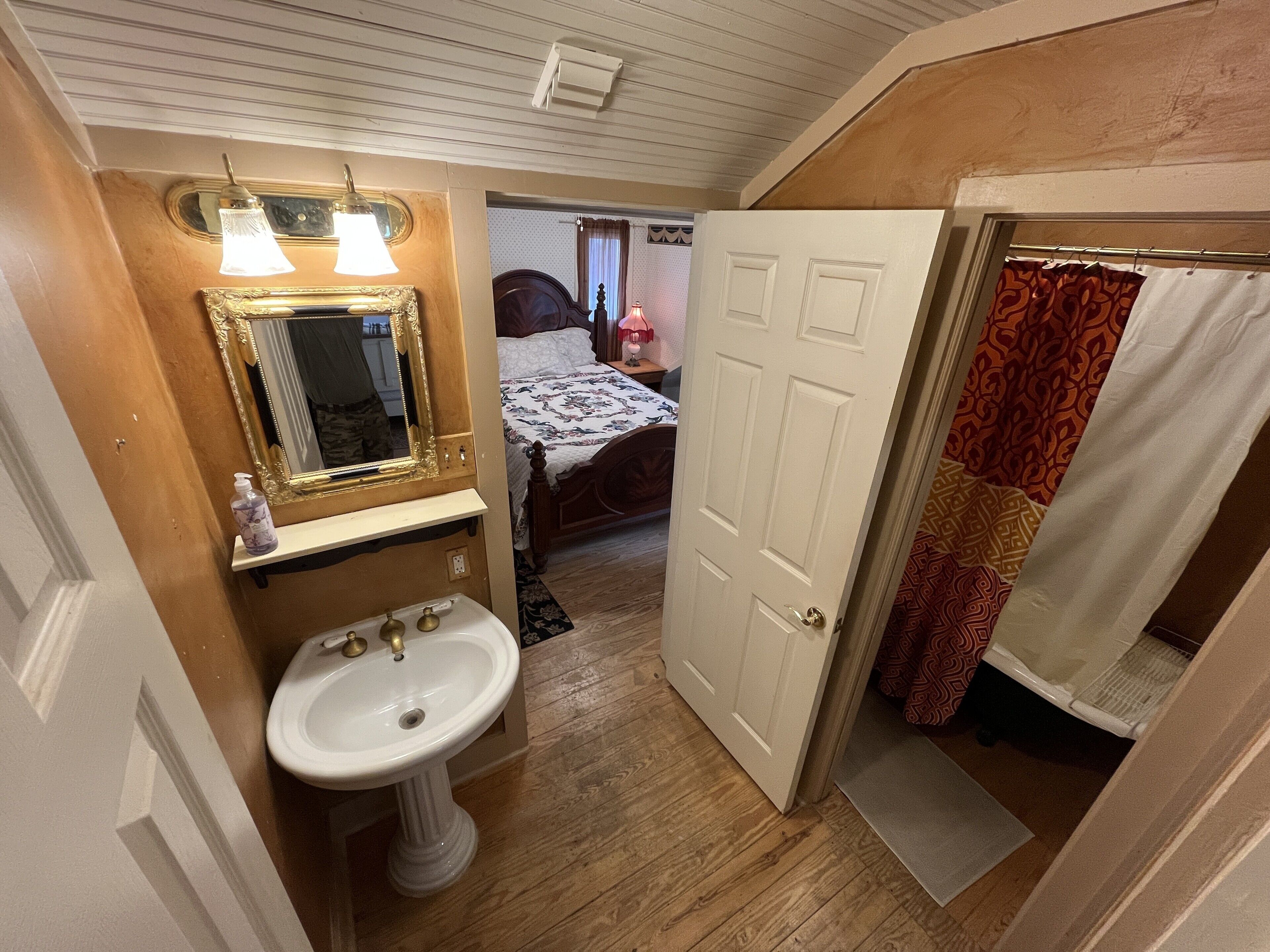 A bathroom with a pedestal sink, a mirror with an ornate frame, and a door leading to a bedroom.