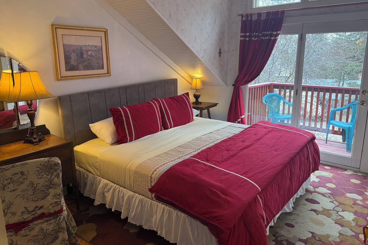 A bedroom with a bed, a nightstand, a lamp, a chair, a balcony with a view, and a painting on the wall.