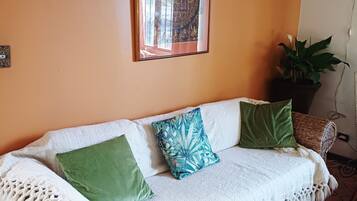 Une pièce chaleureuse avec un canapé blanc en tissu tissé, des coussins à motifs verts et bleus, une œuvre d’art encadrée au mur et une plante en pot.