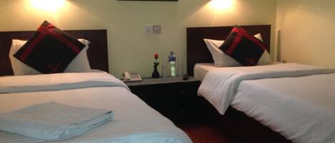 A hotel room with two beds, each with a pillow and a blanket. There is a nightstand with a phone, a vase with a rose, and a water bottle.