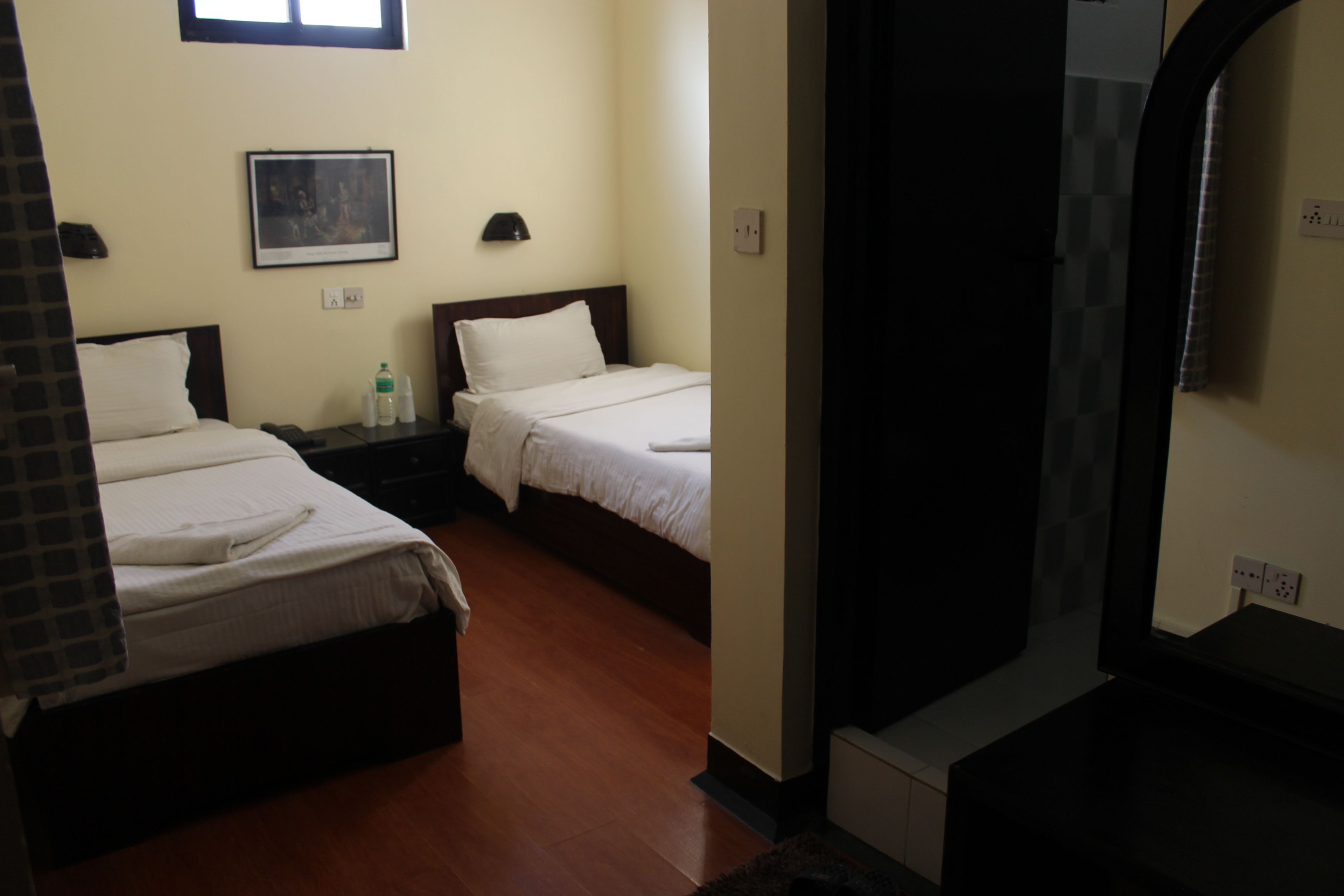 Habitación de hotel con dos camas individuales, una mesita de noche con lámpara, un cuadro en la pared y una ventana.