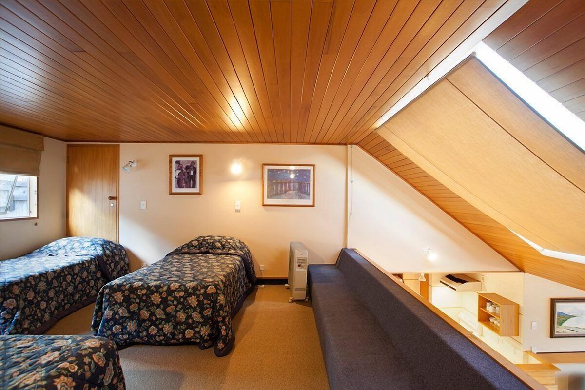 A room with a sloped ceiling, two beds with floral bedding, a bench, a radiator, and framed pictures on the walls.