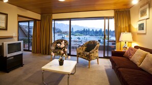 Une chambre d’hôtel dotée d’une grande porte-fenêtre coulissante donnant sur un balcon avec vue sur les montagnes. On y trouve un téléviseur posé sur un meuble, une table basse avec un vase de fleurs et un canapé avec des coussins.