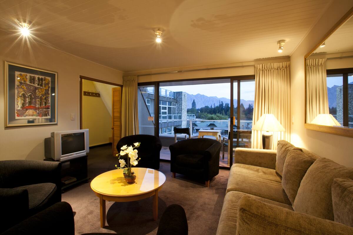 A hotel room with a sofa, armchair, and a small table with a potted plant. There is a TV on a stand and a window with curtains.