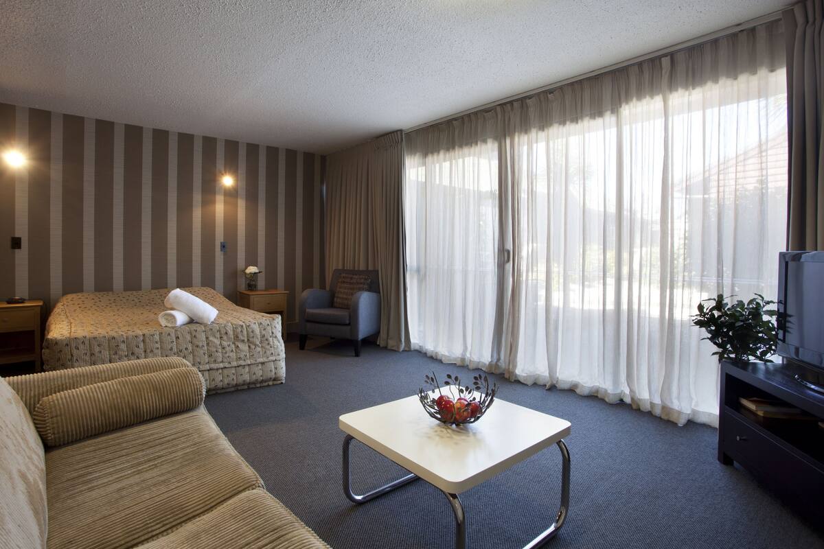 A hotel room with a bed, a sofa, a small table with a fruit bowl, a TV, and curtains.