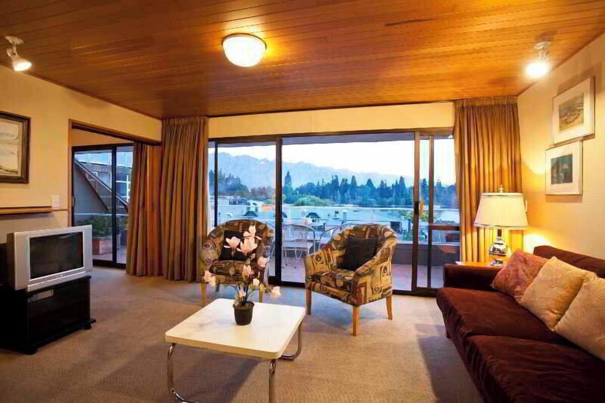 A living room with a TV, two patterned armchairs, a sofa, a coffee table, and a view of mountains through large windows.