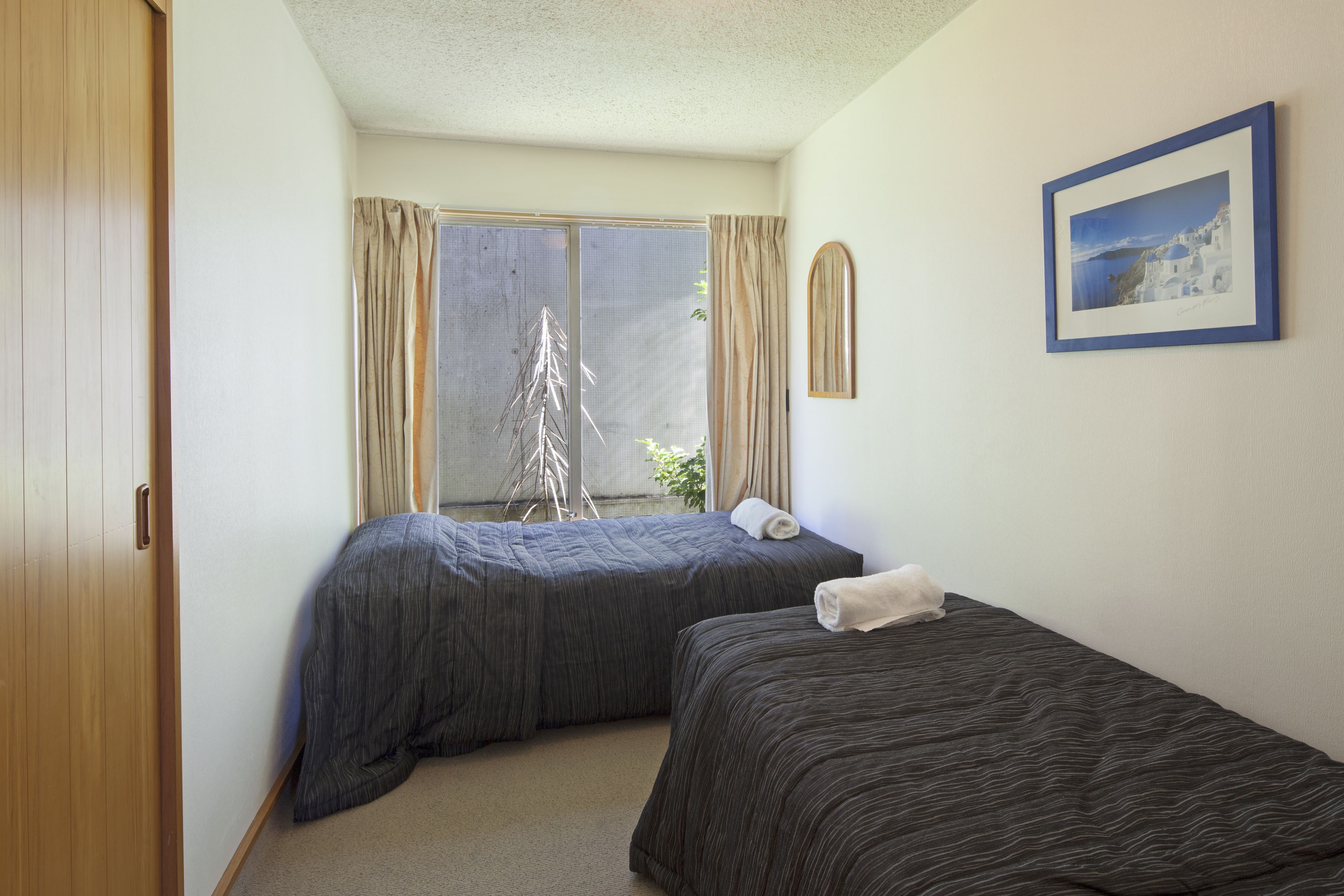 A room with two beds, a window with curtains, a framed picture on the wall, and a wooden door.