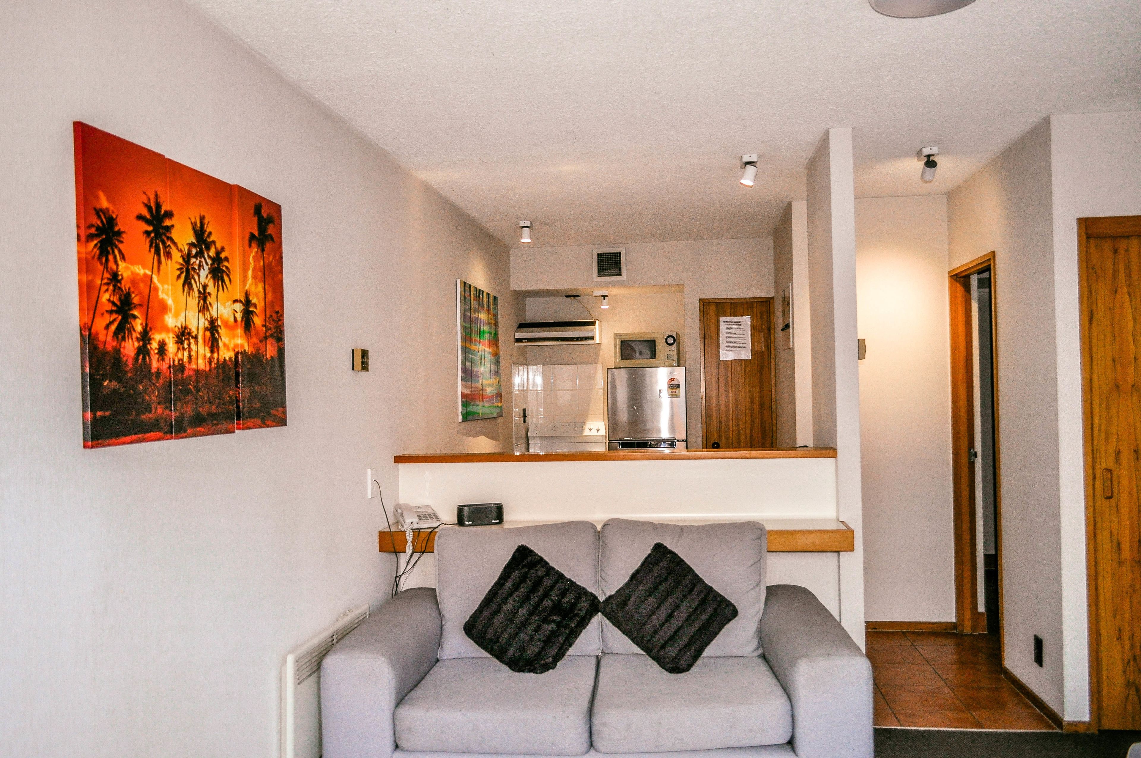 A living room with a grey sofa, two black cushions, a painting of palm trees, and a kitchen in the background.