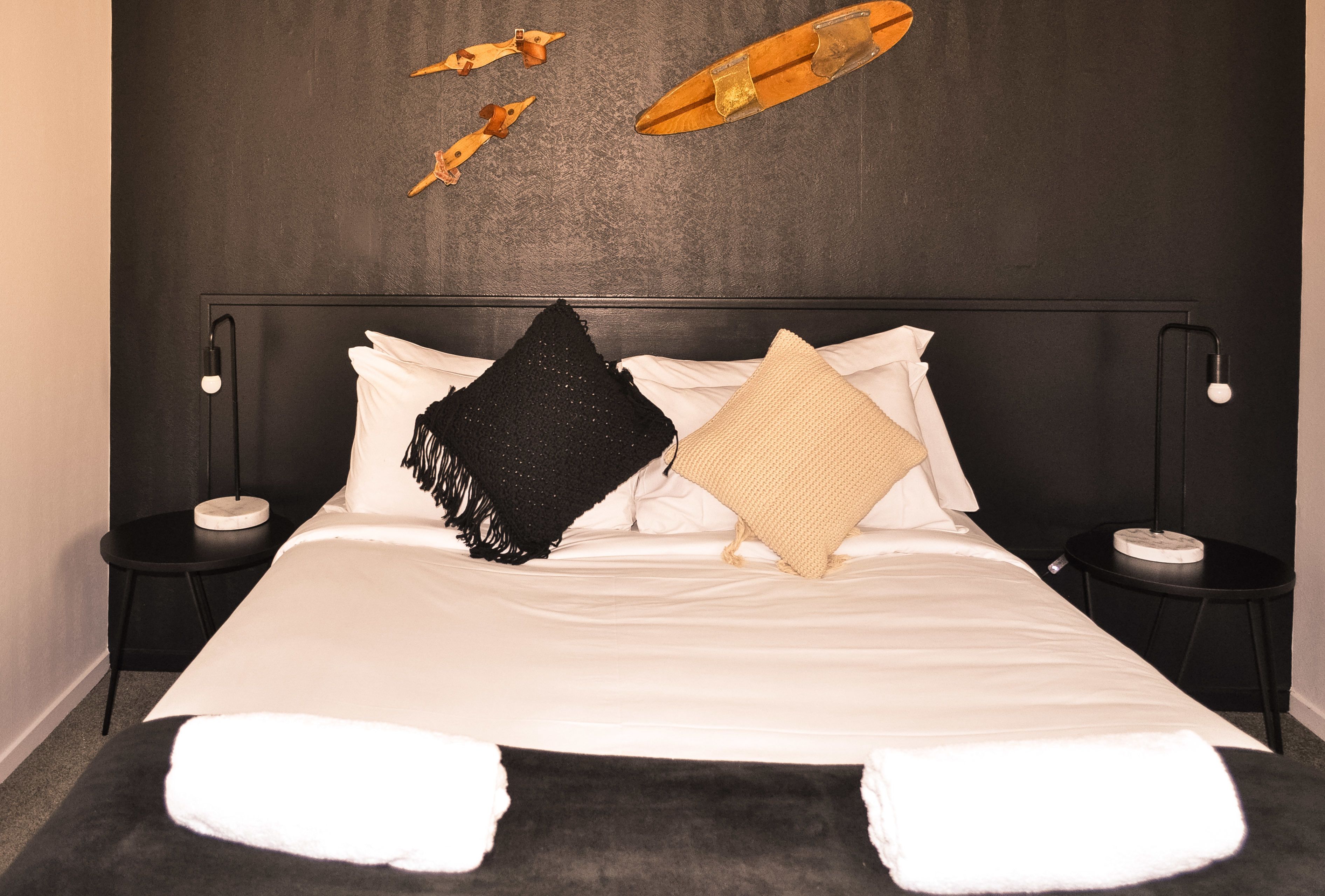 Una cama con ropa de cama blanca, un plaid negro y un plaid beige. Hay dos mesitas de noche con lámparas y una mesita pequeña al lado de la cama.