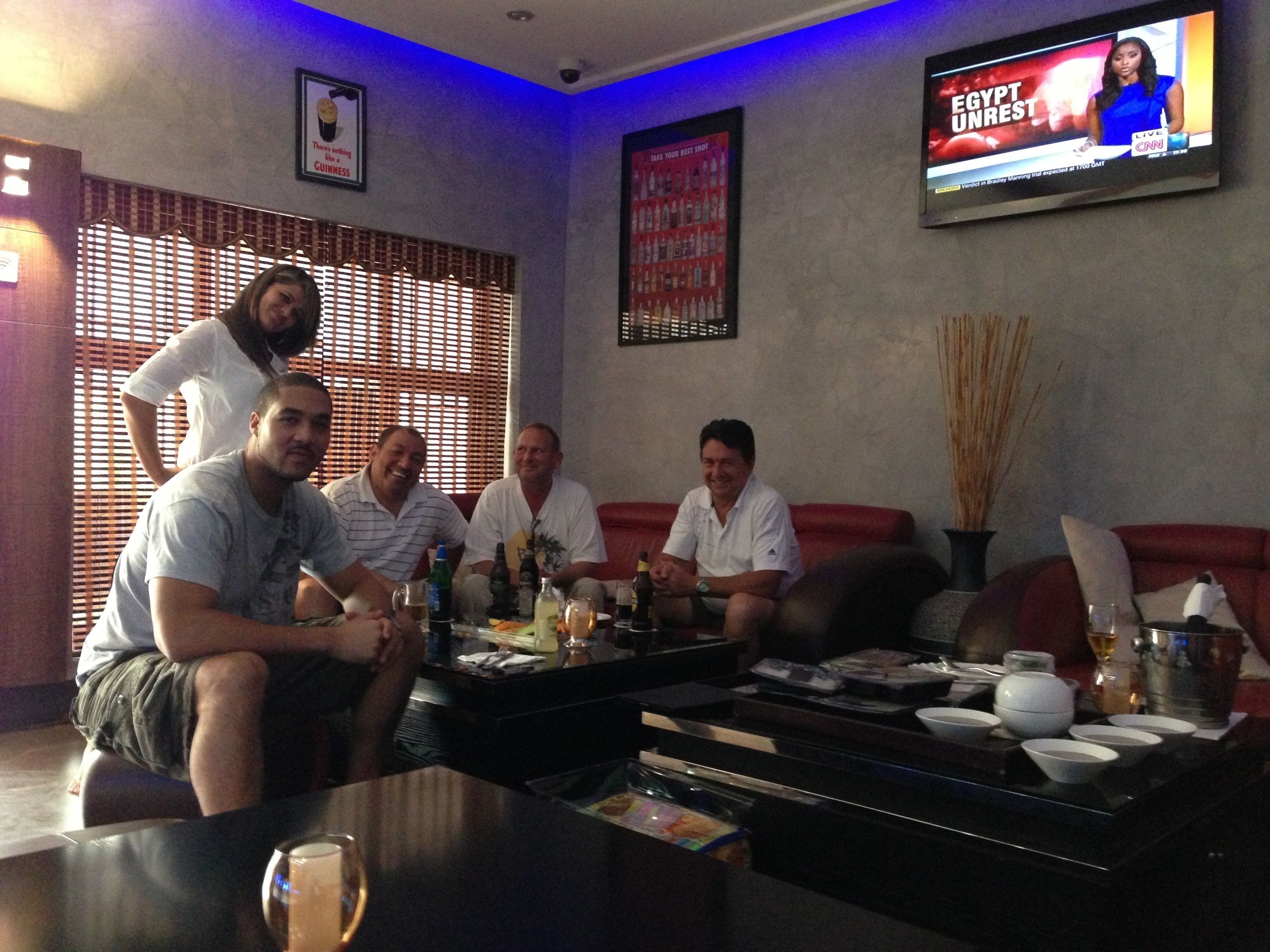 A group of people in a lounge area with a television displaying a news program, a coffee table with various items, and a wall with a Guinness advertisement.