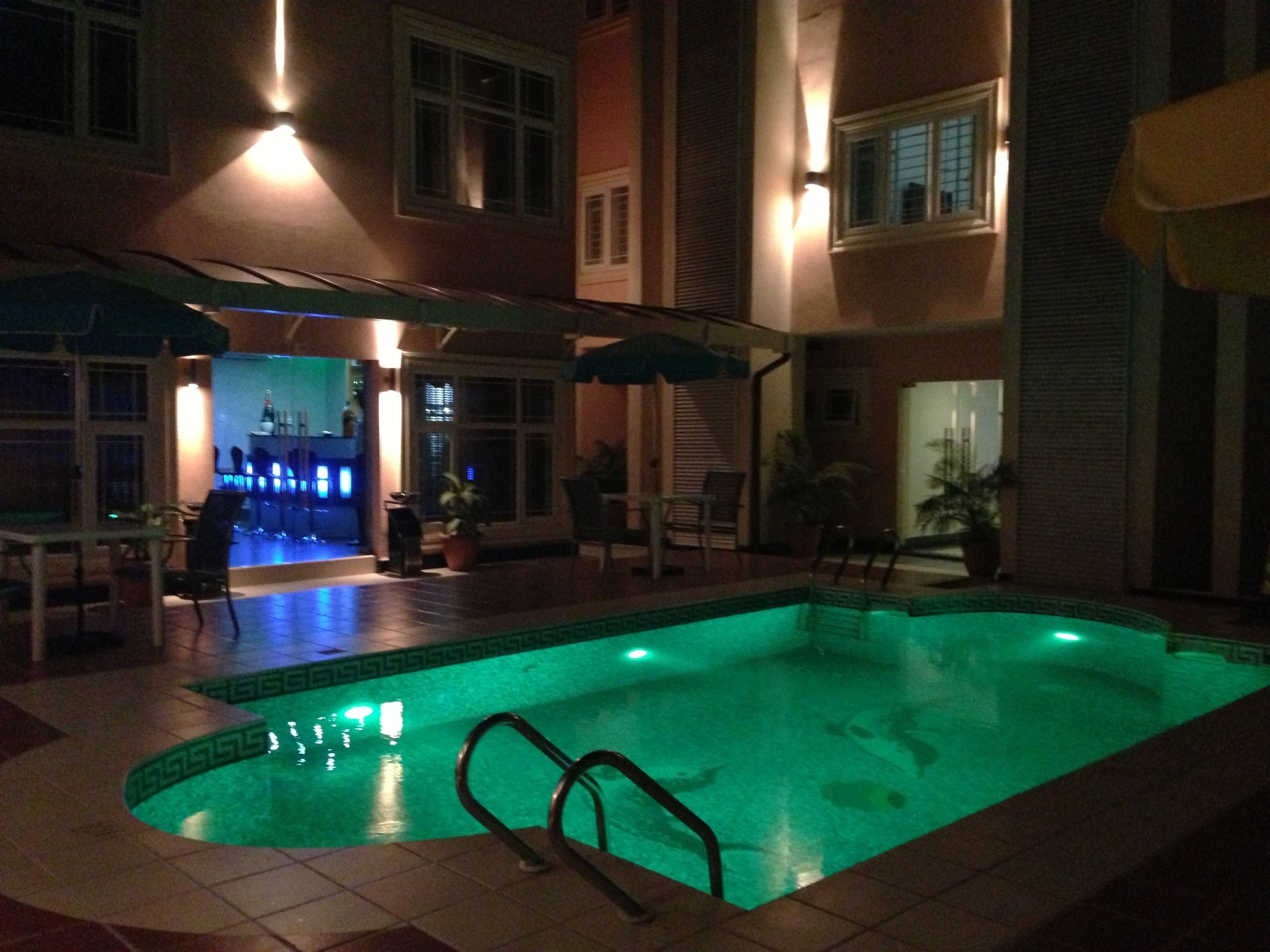 Une piscine intérieure éclairée pour la nuit, dont l’eau a une teinte verdâtre, entourée d’un patio aménagé avec des chaises et des parasols.