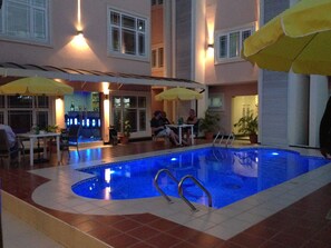 An indoor swimming pool with tiled flooring, yellow umbrellas, and a few people seated around the pool area. - GrandBee Suites (Lagos)
