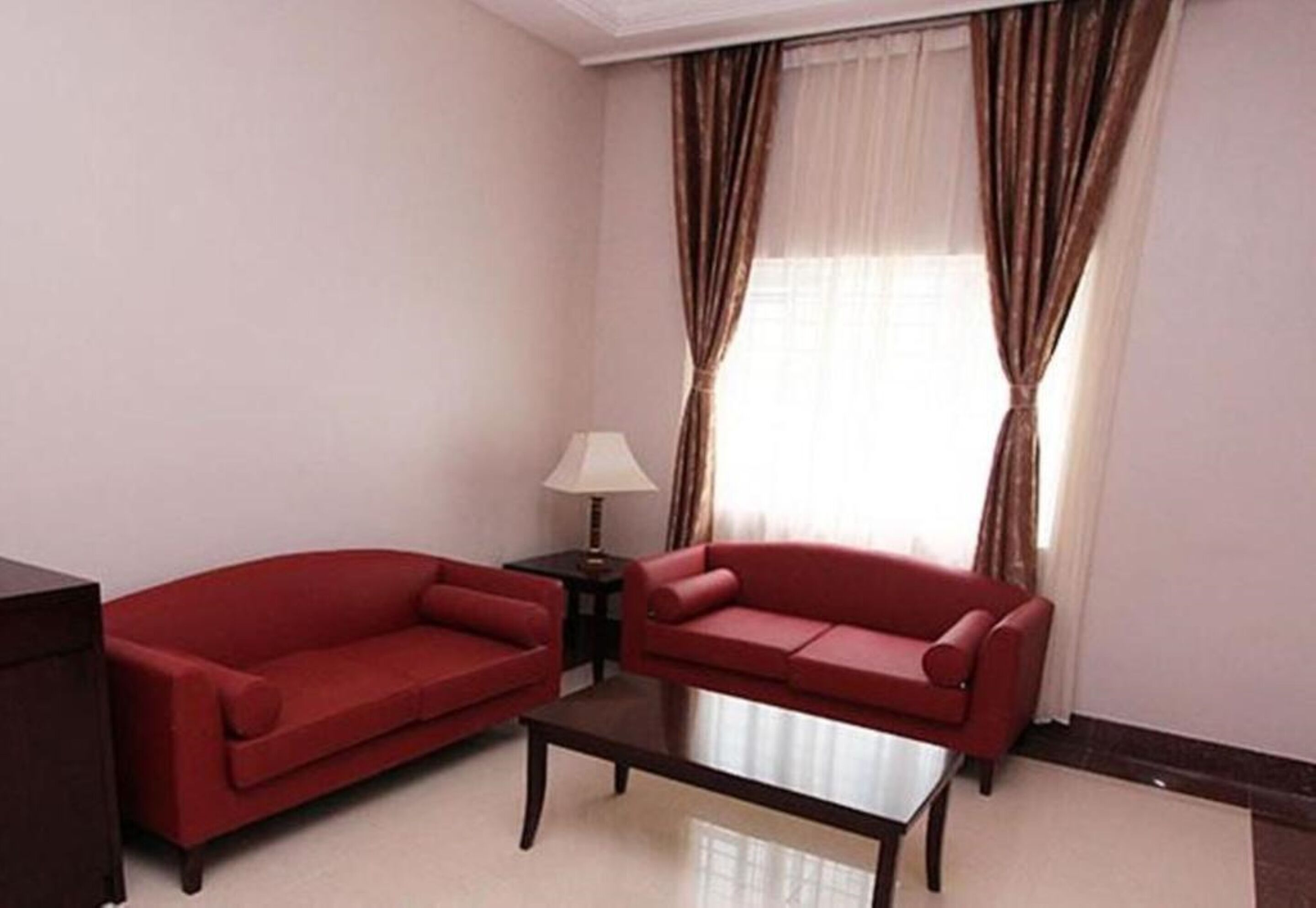 A room with two red sofas, a small table, a lamp, and a window with curtains.