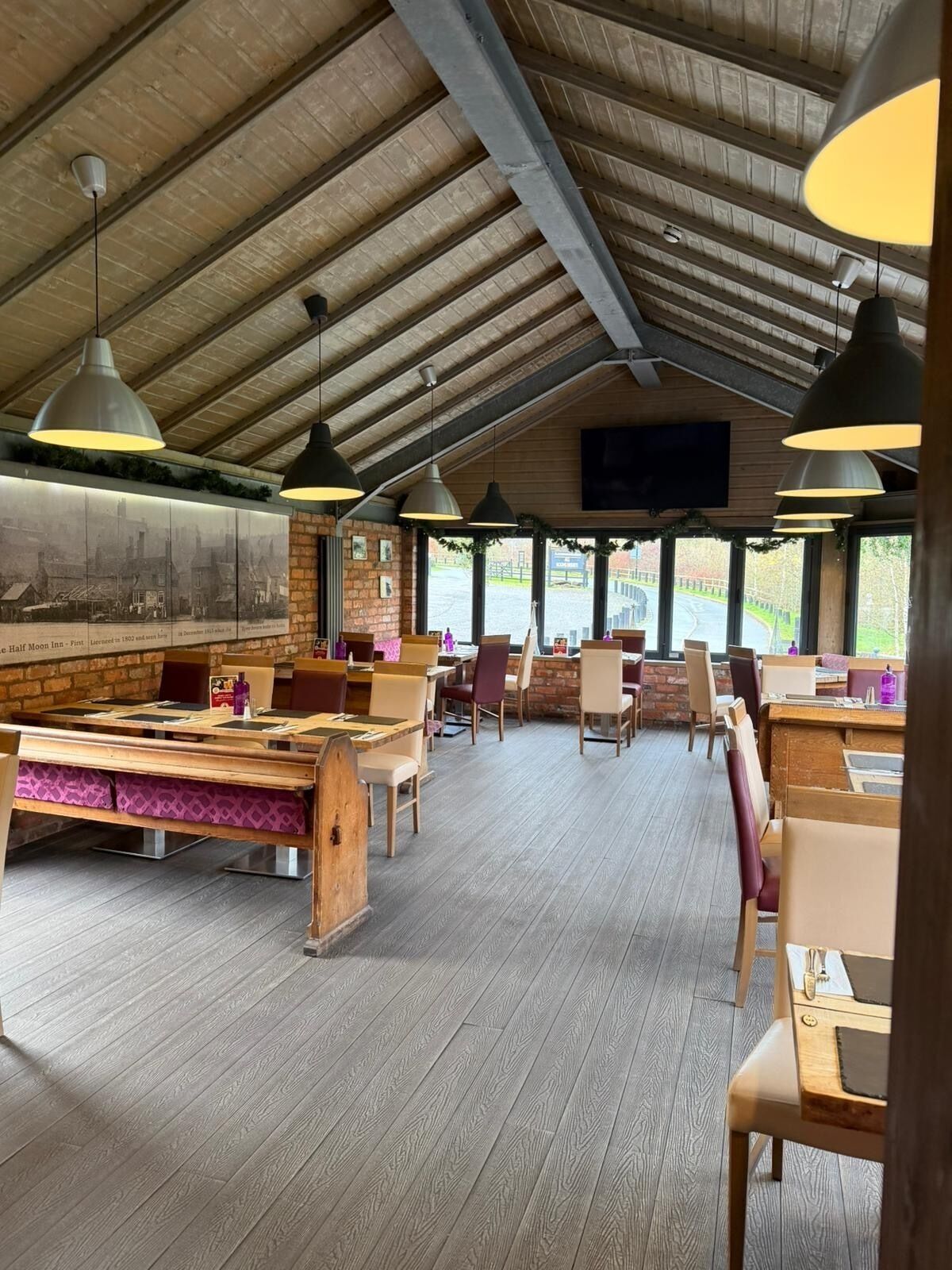 Un restaurante amplio con mesas y sillas de madera, una pared de ladrillo con un mural histórico y ventanas grandes que ofrecen vistas al exterior.