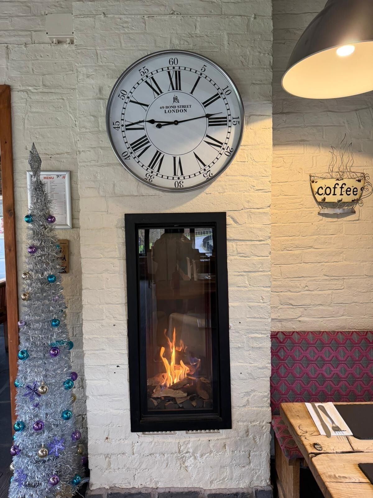 A wall clock with Roman numerals, a framed menu, a decorative Christmas tree, a fireplace, and a coffee cup sign.