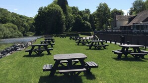 A grassy area with picnic tables arranged in rows, a river in the background, and a building with a chimney. - Half Moon Jackfield (Telford)