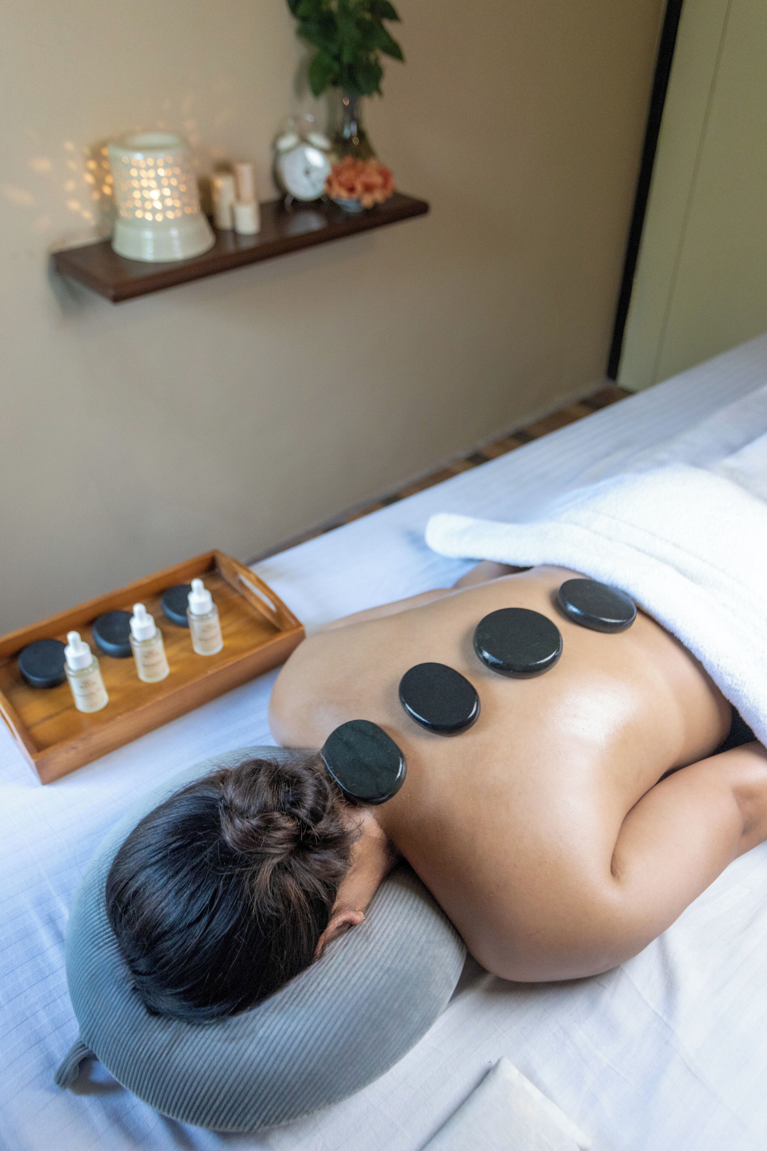 A person receiving a body treatment with multiple black circular stones on their back, a tray with bottles, and a shelf with various items in the background.