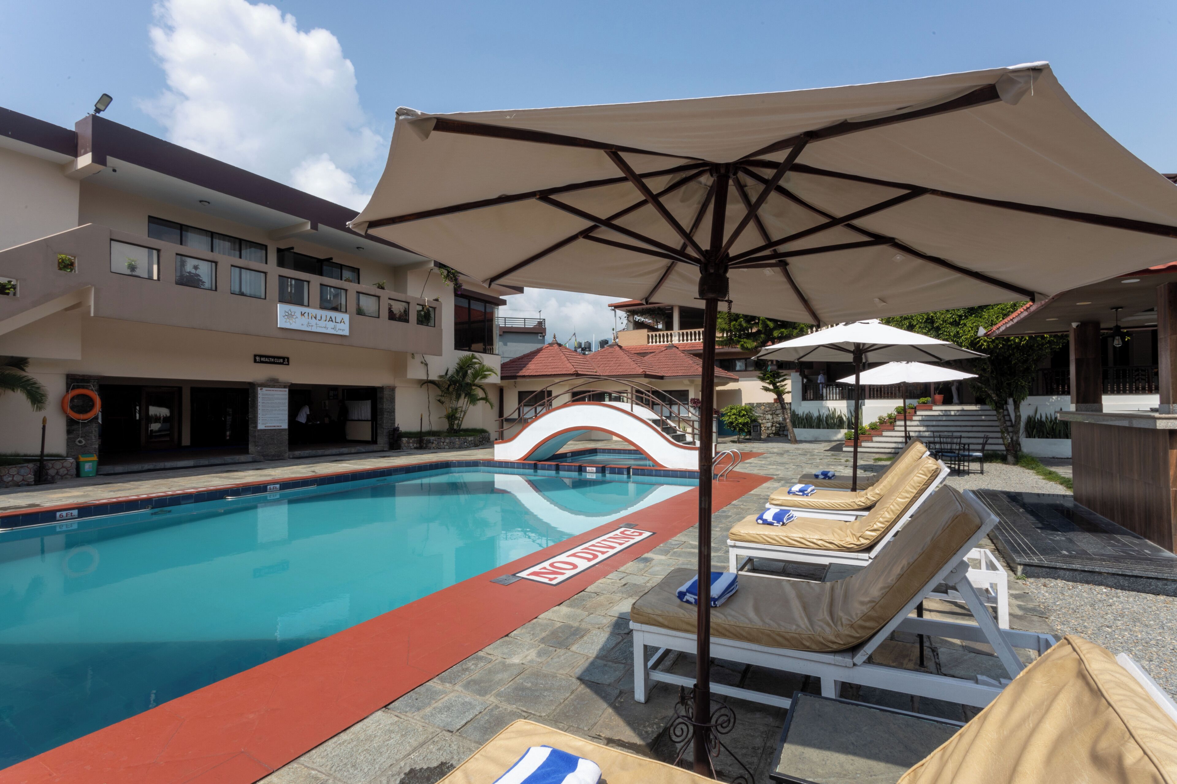 Una piscina con sillas de descanso y sombrillas, un letrero de club de salud y un edificio con un letrero que dice 'KINJALAL'.