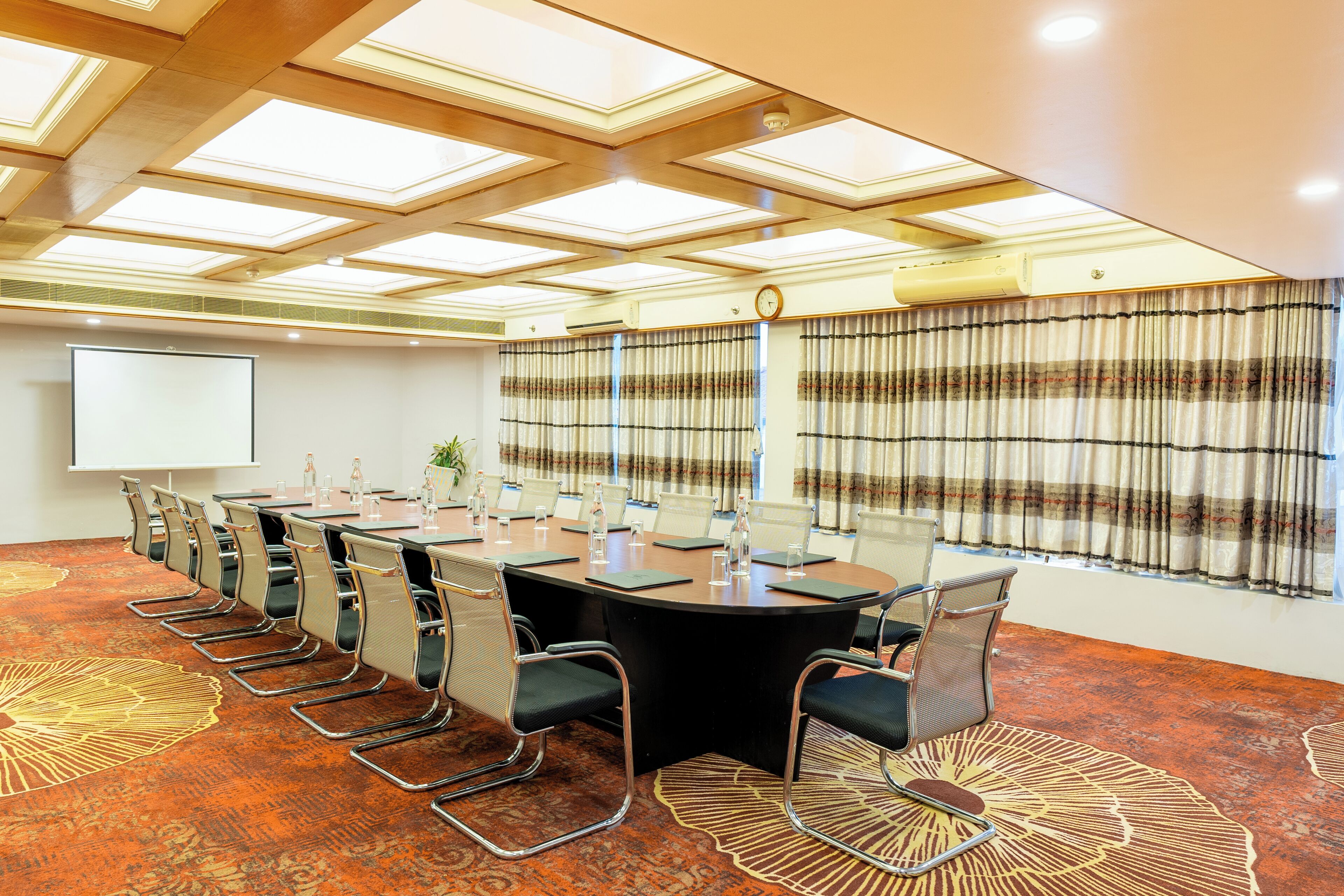 A conference room with a large oval table, multiple chairs, a projector screen, and a patterned carpet.