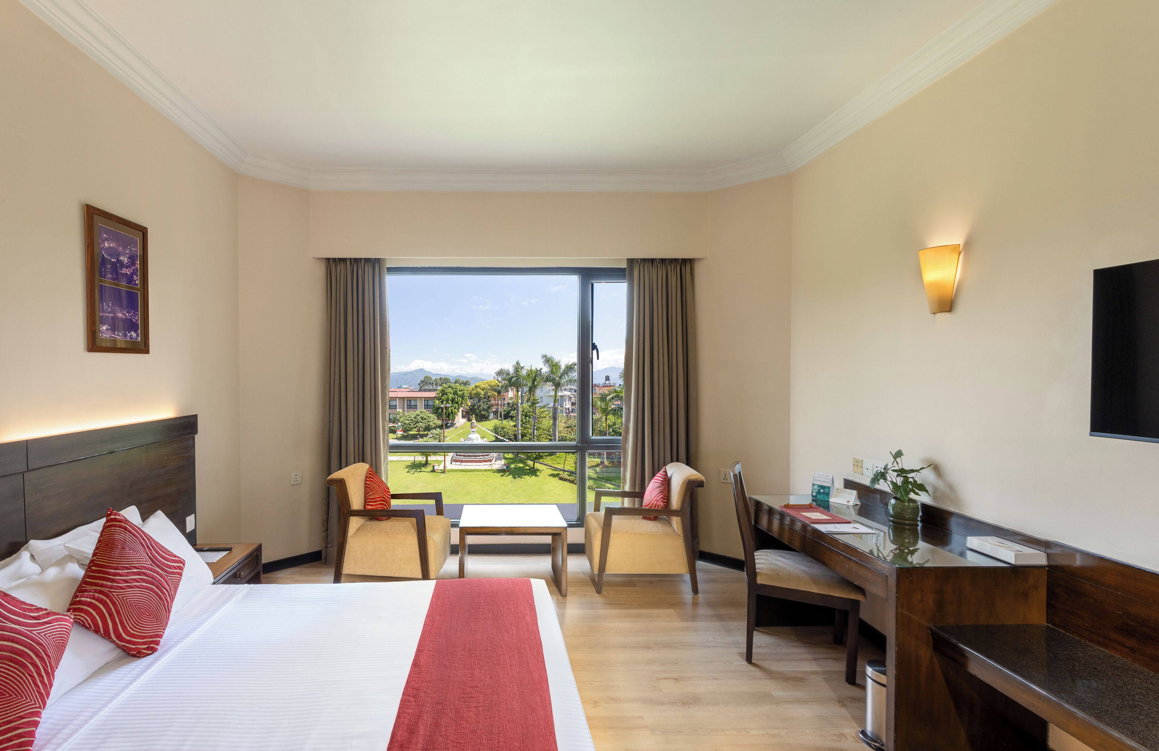 A hotel room with a bed, a desk, a chair, a small table, and a view of a garden and mountains.
