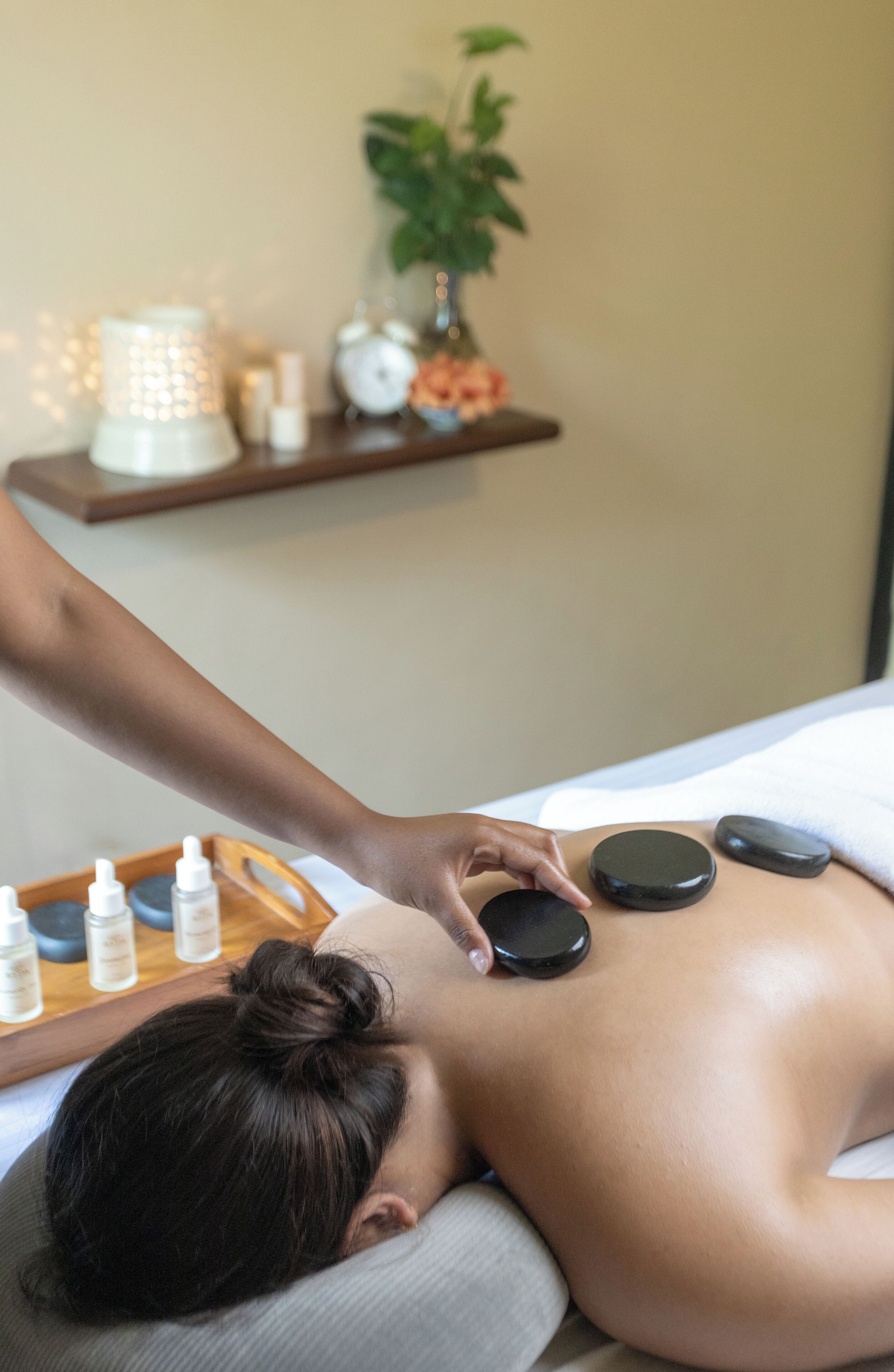 A person receiving a back massage with stones, a plant, and bottles on a wooden tray in a room.